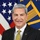 Kelly McKeague A formal official portrait of a middle-aged man with gray-streaked hair wearing a dark suit, white dress shirt, and gold patterned tie. He is smiling and posed in front of an American flag and what appears to be a Department of Defense or military organizational flag. A POW/MIA lapel pin is visible on his jacket.