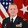 U.S. Army Brig. Gen. Joseph W. Rank An official U.S. Army military portrait of an officer identified by nameplate as 'RANK', wearing the Army Service Uniform (ASU) in dress blue. The subject is posed in front of a U.S. national flag and a red general officer flag displaying white stars. The uniform displays numerous medals, ribbons, badges, and insignia consistent with a senior officer.