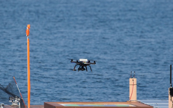 A quadcopter drone hovers above a floating platform or barge on open water. The platform appears to be equipped with a drone landing pad (the raised wooden-framed square structure), communication antennas, and an orange wind indicator pole. This setup suggests an autonomous or remote drone docking/landing station deployed on water.