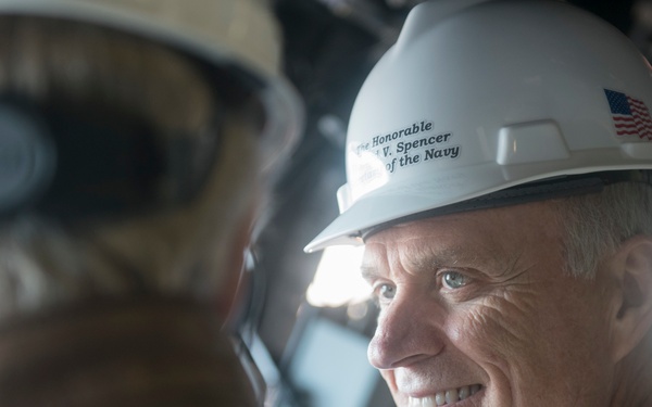 A man wearing a white hard hat labeled 'The Honorable [Richard] V. Spencer, Secretary of the Navy' with an American flag decal is smiling during what appears to be an official naval facility visit. He is engaged in conversation with another individual whose back is to the camera. The setting appears to be an industrial or shipyard environment.