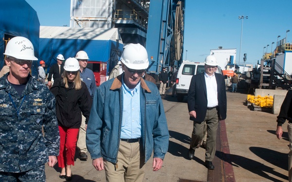 A group of individuals wearing white safety hard hats are walking through what appears to be a naval shipyard or industrial port facility. A U.S. Navy sailor in digital camouflage uniform with a name tape reading 'BAUER' leads the group alongside a civilian in a denim jacket. The setting suggests an official tour or inspection of a shipbuilding or repair facility.