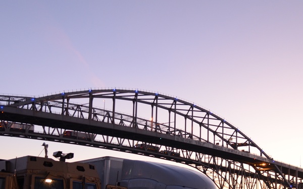 A dusk photograph showing two large trucks parked near a waterfront with a large steel arch bridge illuminated by blue lights in the background. The scene captures a sunset sky with warm orange and pink hues reflecting off the water. One truck appears to be a military-style tan/yellow vehicle while the other is a modern white commercial semi-truck.