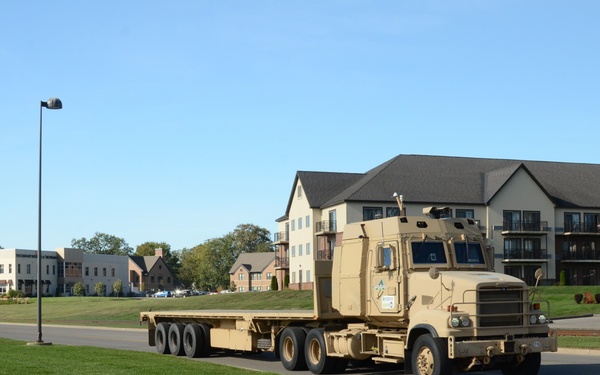 A tan/desert-colored military flatbed semi-truck is driving along a road through what appears to be a military installation or base housing area. The truck, consistent with a Heavy Expanded Mobility Tactical Truck (HEMTT) or similar military transport vehicle, has an empty flatbed trailer. The surroundings include well-maintained grass, residential-style buildings, street lights, and a clear blue sky.