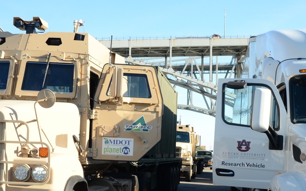 The image shows a convoy of military and research vehicles parked or staged in what appears to be a parking area or staging ground near an overpass or bridge structure. The vehicles include tan/beige military trucks and a white Auburn University Research Vehicle, with visible sponsorship logos from MDOT and Planet M on the military vehicles. This appears to be a demonstration or testing event involving autonomous or connected vehicle technology.