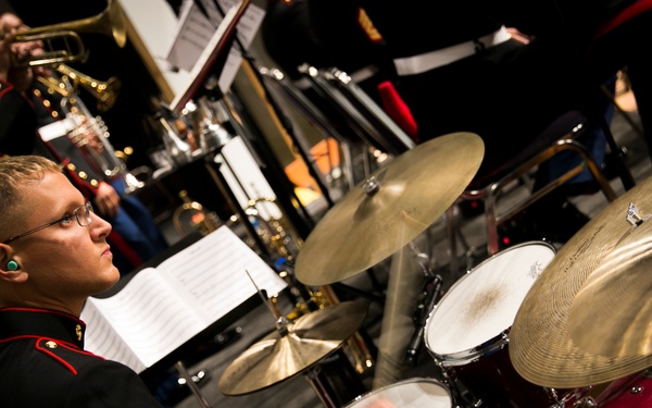 A U.S. Marine Corps musician in dress blues uniform plays a drum kit during a formal band performance. The drummer, wearing the rank insignia of a Corporal, is captured mid-play with motion blur on his hands. Behind him, other Marines in dress uniforms play brass instruments with sheet music on stands.