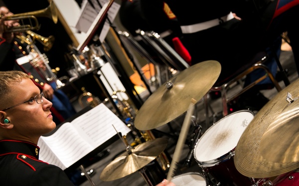 A U.S. Marine Corps musician in dress blues uniform plays a drum kit during a formal band performance. The drummer, wearing glasses and an in-ear monitor, bears the rank insignia of a Corporal on his sleeve. Other Marine band members are visible in the background playing brass instruments.