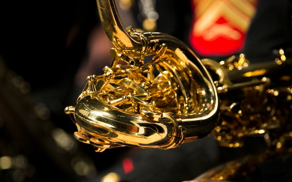 Close-up photograph of U.S. Marine Corps musicians holding brass instruments, likely French horns or similar brass instruments. The musicians are wearing formal dress blue uniforms with gold buttons and red trim, characteristic of the United States Marine Corps Band. The shallow depth of field emphasizes the gleaming gold instruments in the foreground.
