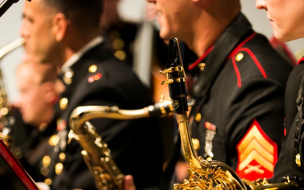 A group of United States Marine Corps musicians are seated and performing in a military band concert. They are wearing the distinctive dress blue uniform with red trim and gold buttons. Multiple brass and woodwind instruments are visible, including saxophones and a French horn.