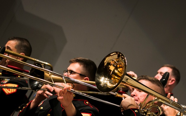 A group of United States Marine Corps musicians are performing in a formal concert setting. The musicians are dressed in their distinctive dress blue uniforms with red trim and gold buttons, playing brass instruments. The image captures a close-up view of the trombone and brass section mid-performance.