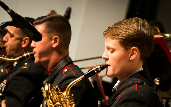 A group of United States Marine Corps musicians performing in a formal band concert, wearing dress blues uniforms with red trim and gold buttons. The musicians are seated and playing brass and woodwind instruments while reading from red music stands. The foreground features a sergeant identifiable by the chevron rank insignia on the sleeve.