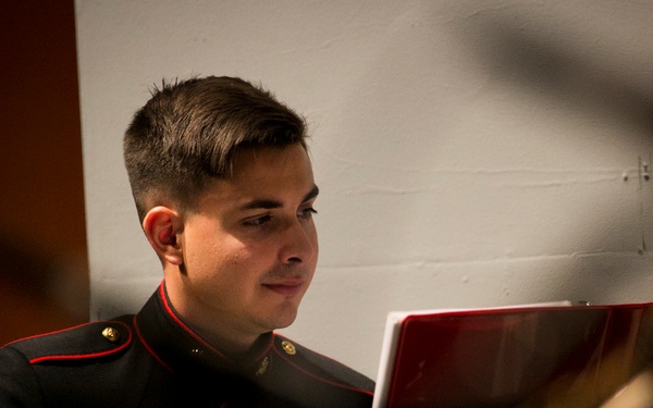 A young male musician in a U.S. Marine Corps dress uniform is seated and reading sheet music from a red music folder on a stand. He appears to be part of a military band ensemble, with other musicians and music stands visible in the blurred foreground. The image is taken at close range with a shallow depth of field, keeping the subject in sharp focus.