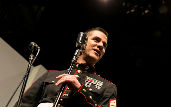 A male individual wearing a United States Marine Corps dress blue uniform is standing at a vintage-style microphone on a stage. He appears to be performing or speaking at a formal event, with stage lighting visible overhead. The photo is taken from a low angle, emphasizing the subject's presence and uniform details.