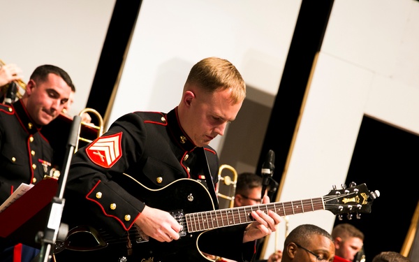 A group of United States Marine Corps musicians perform in formal dress blues uniforms during what appears to be an official concert or ceremony. The central figure is a sergeant playing a black Guild hollow-body electric guitar, surrounded by fellow Marines playing brass instruments including trumpet and trombone. The ensemble appears to be a Marine Corps jazz or big band ensemble, with music stands visible throughout.