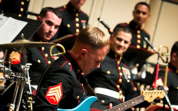 A group of U.S. Marine Corps musicians are performing in a formal concert setting, all dressed in their distinctive dress blue uniforms with gold buttons and red trim. The foreground features a sergeant playing a blue Fender Stratocaster electric guitar with focused intensity. Behind him, other Marines are visible with drums, brass instruments, and music stands.