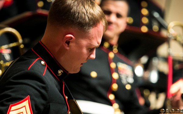 A U.S. Marine Corps sergeant is playing a blue Fender Stratocaster electric guitar during what appears to be a formal military band performance. He is wearing the dress blues uniform with a red chevron sergeant rank insignia on his sleeve. Several other Marines in dress blues are visible in the background, some holding brass instruments.