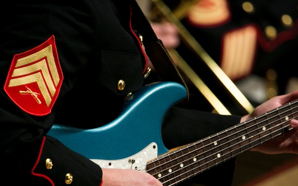 A military musician in formal dress uniform plays a blue electric guitar, identifiable as a Fender Stratocaster-style instrument. The uniform features a red and gold sergeant's chevron patch with crossed rifles, consistent with U.S. Marine Corps dress blues. Additional uniformed musicians are visible in the background, suggesting a formal military band performance.