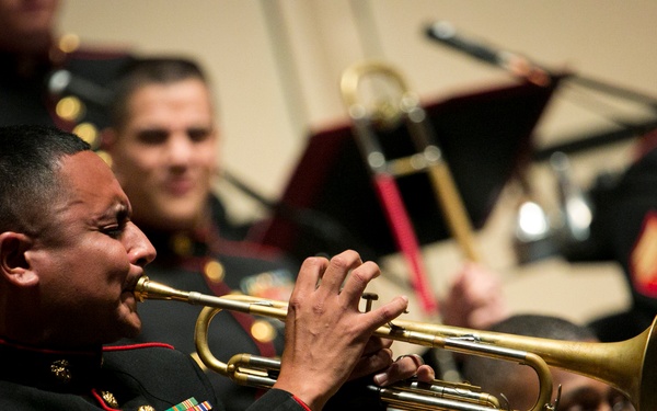 A group of United States Marine Corps musicians are performing in a formal band concert. The foreground features a Marine sergeant playing a brass trumpet while wearing the distinctive black and red dress uniform. Multiple other Marines with various instruments are visible in the background.