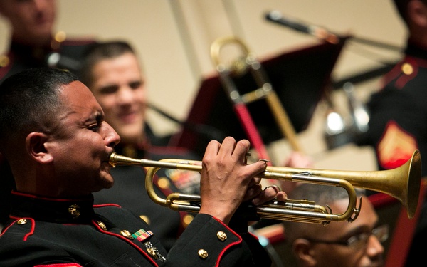 A United States Marine Corps band is performing at a formal concert. The foreground features a Marine sergeant playing a brass trumpet while wearing the distinctive dress blues uniform. Multiple other Marines in the same formal attire are visible in the background, some holding instruments.