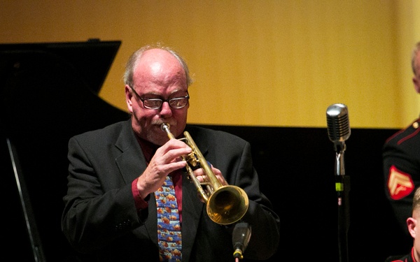A performance featuring a civilian trumpeter playing a brass trumpet at center stage, accompanied by at least two U.S. Marine Corps musicians in dress uniform. A vintage-style microphone is positioned near the trumpet player, and a grand piano is visible on the left side of the frame. The setting appears to be a formal concert hall or performance venue.