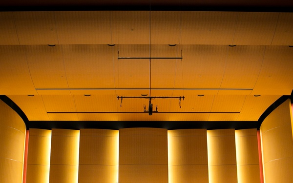 A large ensemble, appearing to be a military band based on the uniforms visible, performs on a concert hall stage with warm golden acoustic paneling. The musicians are arranged in a big band or jazz ensemble configuration with brass, woodwind, and rhythm section instruments. An audience is visible in silhouette in the foreground, watching the performance.