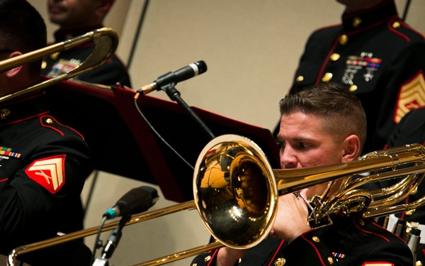 A group of United States Marine Corps musicians perform in a formal concert setting, wearing dress blues uniforms with gold buttons and red trim. The foreground features a Marine playing a valve trombone, with additional trombone players visible behind him. Music stands with red folders are positioned in front of the performers.
