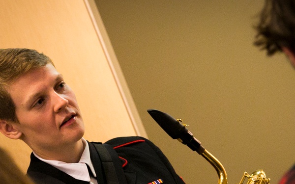 A person in a dark military dress uniform with medals and ribbons is holding a brass saxophone and appears to be speaking or demonstrating to others nearby. The setting appears to be an indoor venue, possibly during a meet-and-greet or educational outreach event. Two other individuals are partially visible in the foreground and right side of the frame.