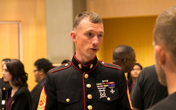 A U.S. Marine in dress blues uniform is engaged in conversation with a group of civilians in what appears to be an indoor venue or event hall. The Marine displays sergeant major rank insignia and several service ribbons and medals on his uniform. The setting suggests a recruiting event, community outreach, or formal military engagement.