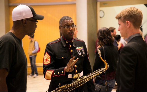 A U.S. Marine in dress blues uniform, holding a saxophone, is engaged in conversation with two civilians in what appears to be a building lobby or hallway. The Marine displays multiple service ribbons and chevrons indicating a senior non-commissioned officer rank (Master Sergeant). The setting suggests a post-performance or public engagement event.
