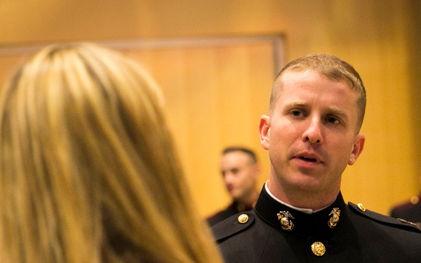 A U.S. Marine Corps officer in dress blues uniform is engaged in conversation with two women in a formal indoor setting. The officer displays several ribbons and medals on his chest, suggesting a ceremonial or formal military event. Additional Marines in uniform are visible in the background.