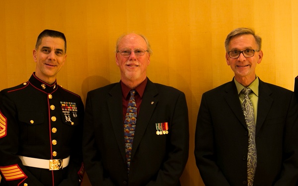 Five men pose for a formal group photograph against a wood-paneled wall. Three individuals are wearing U.S. Marine Corps dress blue uniforms with gold buttons, red trim, white belts, and visible medals/ribbons, while two men in civilian formal attire also display miniature medal ribbons on their lapels. The setting appears to be an indoor formal military or ceremonial event.