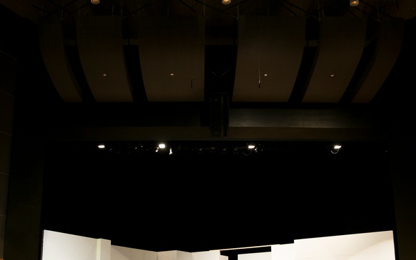 A military band is performing on a well-lit stage in what appears to be a formal auditorium or concert hall. The musicians are dressed in military uniforms and are arranged in a big band or jazz ensemble formation with music stands. The audience is visible in silhouette in the foreground, watching the performance.