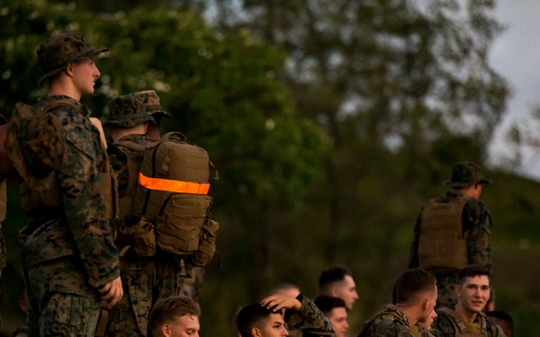 A group of military personnel in woodland camouflage uniforms are gathered on a grassy area, with some standing and others seated on the ground. They are equipped with tactical backpacks, some marked with high-visibility orange and yellow reflective strips. The setting appears to be an outdoor training or assembly area during low-light conditions, likely dawn or dusk.