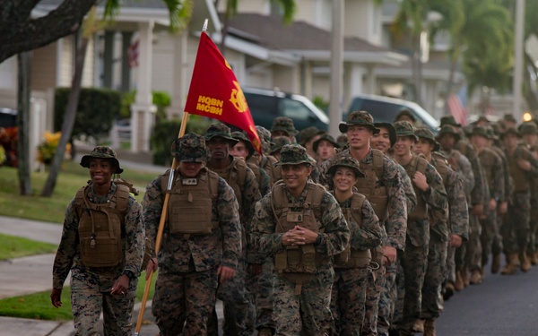 A large column of U.S. Marines in woodland MARPAT camouflage uniforms and plate carriers marches in formation along a residential street, likely on a military base. A guidon bearer carries a red Marine Corps unit flag near the front of the formation. The setting appears to be a base housing area with palm trees and residential homes visible in the background.