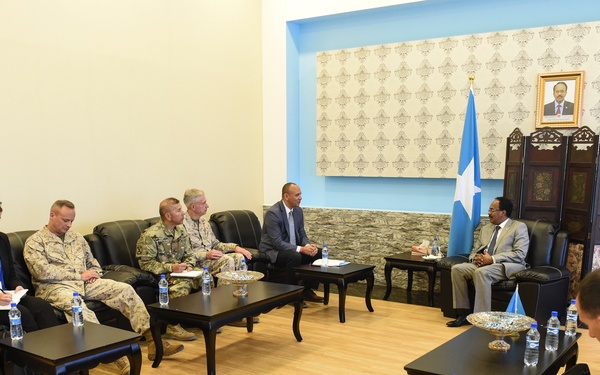 A formal diplomatic or military meeting is taking place in what appears to be an official reception room. Multiple U.S. military personnel in uniform and civilian officials are seated across from a host official, with a Somali flag and a framed portrait visible on the wall, suggesting this is likely a meeting in Somalia. Participants on both sides are taking notes, indicating a structured briefing or negotiation session.