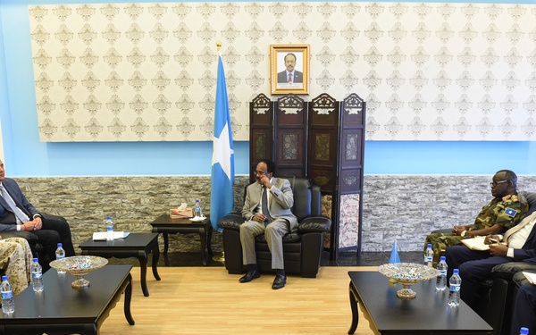 A formal diplomatic or official meeting is taking place in what appears to be a government reception room. A central figure seated in a prominent chair faces two groups arranged on either side, with a Somali flag and a framed portrait visible in the background. The setting, flag, and mixed civilian and military attendees suggest a high-level governmental meeting in Somalia.