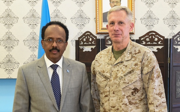 Two individuals pose for a formal photograph in what appears to be an official office setting. One person is wearing a light gray suit with a blue patterned tie and a flag pin on the lapel, while the other is wearing U.S. Marines desert camouflage uniform with a name tape reading 'WALDHAUSER'. A light blue flag is partially visible in the background along with a framed portrait on the wall.