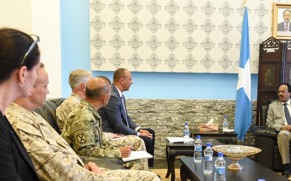 A formal diplomatic or military meeting is taking place in an official reception room. A group of civilians and military personnel in various uniforms are seated facing a man in a suit who appears to be a host official, with a national flag and a framed portrait visible behind him. The setting suggests a high-level governmental or diplomatic briefing.