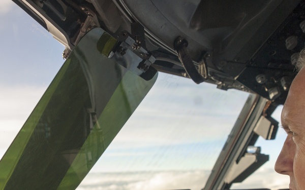 A military pilot is seated in the cockpit of a large aircraft in flight, looking forward through the windscreen at a partly cloudy sky. The cockpit features modern glass cockpit avionics with multiple digital displays visible. The pilot is wearing a flight suit, aviation headset with boom microphone, and is positioned in what appears to be the left (captain's) seat.