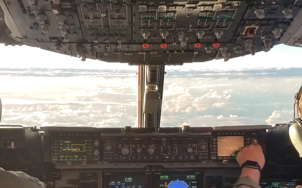 Two military aviators are seated in the cockpit of a large military aircraft, likely a C-17 Globemaster III or similar transport aircraft, flying above a cloud layer. Both crew members are wearing olive drab flight suits and aviation headsets. The cockpit displays multiple illuminated digital instruments and avionics panels, with a clear view of clouds and sky through the windscreen.