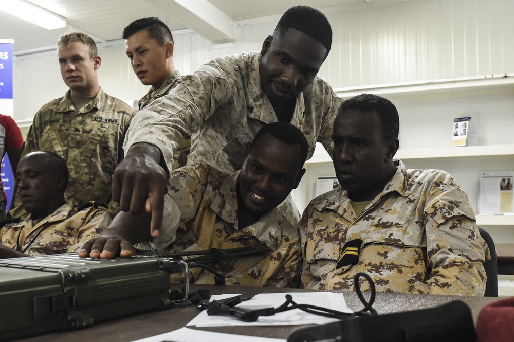 The image shows a group of military personnel from what appears to be multiple nations gathered around a table with military equipment, likely during a training or advisory session. A standing soldier in desert camouflage is pointing at a military-grade laptop or communications device while others observe. The setting appears to be an indoor military facility or classroom environment.
