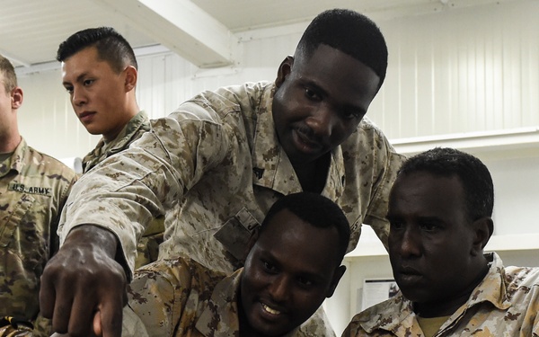 The image shows a group of military personnel from what appears to be multiple nations gathered around a table with military equipment, likely during a training or advisory session. A standing soldier in desert camouflage is pointing at a military-grade laptop or communications device while others observe. The setting appears to be an indoor military facility or classroom environment.