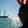 A large historic tall ship with three masts is sailing on open water, viewed from another vessel. Observers in the foreground are silhouetted against the bright sunlight reflecting off the water. An American flag is partially visible in the upper right corner, and a smaller American flag can be seen flying from the tall ship.