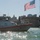 A U.S. Coast Guard vessel numbered 45659 patrols a harbor with an armed personnel standing on deck. An American flag flies prominently in the background, alongside what appears to be a historic tall ship docked at a pier. A city skyline with a distinctive clock tower is visible in the background, consistent with Boston Harbor.