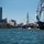A historic tall ship decorated with international signal flags is prominently visible on the right side of the frame, sailing in Boston Harbor. The Boston skyline is visible in the background, including the distinctive Zakim Bridge (Leonard P. Zakim Bunker Hill Memorial Bridge) and TD Garden arena. A U.S. Coast Guard vessel is also visible escorting or patrolling nearby.
