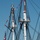 The image shows the upper masts and rigging of a large tall ship, likely a historic naval vessel, decorated with colorful signal flags. Crew members dressed in white uniforms are stationed on the crow's nest platforms of multiple masts. An American flag is visible in the lower left, suggesting this may be a ceremonial or public event.