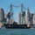A large historic tall ship, decorated with colorful signal flags strung between its masts and flying an American flag, sails in a harbor with a major city skyline in the background. A red tugboat labeled 'Vincent D. Tibbetts' accompanies the vessel. The scene appears to be a ceremonial or commemorative sailing event in Boston Harbor, with the distinctive Custom House Tower visible on the right.
