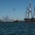 A large historic three-masted sailing vessel, consistent in appearance with USS Constitution, is underway in a harbor with a city skyline visible in the background. The ship is decorated with colorful signal flags strung between its masts and flies an American flag. A fireboat is spraying water jets in celebration, accompanied by a tugboat and a Coast Guard patrol vessel.