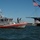 A U.S. Coast Guard vessel (hull number 45659) is patrolling near a large historic tall ship flying an American flag. A Coast Guard personnel member is standing on the bow of the vessel, appearing to man a mounted weapon. An aircraft is visible in the background sky, and the scene appears to take place in a harbor or bay.