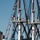 The image shows the upper masts and rigging of a tall ship decorated with numerous international signal flags. Crew members dressed in white uniforms are stationed on the crow's nest platforms at multiple mast levels. A modern brick building is visible in the background, suggesting the ship is docked in an urban harbor.