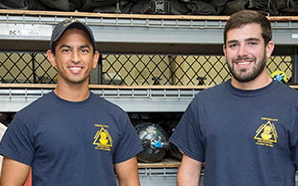 Two young men are standing together and smiling in what appears to be a fire station equipment storage area. Both are wearing matching navy blue t-shirts with a yellow triangular logo on the chest. The setting suggests they may be firefighters or fire academy trainees.